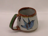 Ken Edwards Truncated Mug 3.5"x5.5" (T7) with brown rim.   It is natural grey clay color background with birds, butterflies, and leaves in blue, green, black and brown on the outside.