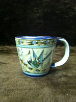 Ken Edwards Collection coffee mug with blue rim.   It is natural grey clay color background with birds, butterflies, and leaves in blue, green, black and brown on the outside.