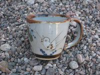 Ken Edwards Mug with brown rim and handle.   It is natural grey clay color background with birds, butterflies, and leaves in blue, green, black and brown on the outside.