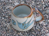 Ken Edwards Truncated Mug 3.5"x5.5" (T7) with brown rim. It is natural grey clay color background with birds, butterflies, and leaves in blue, green, black and brown on the outside.