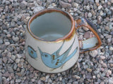 Ken Edwards Truncated Mug 3.5"x5.5" (T7) with brown rim. It is natural grey clay color background with birds, butterflies, and leaves in blue, green, black and brown on the outside.