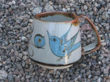 Ken Edwards Truncated Mug 3.5"x5.5" (T7) with brown rim. It is natural grey clay color background with birds, butterflies, and leaves in blue, green, black and brown on the outside.